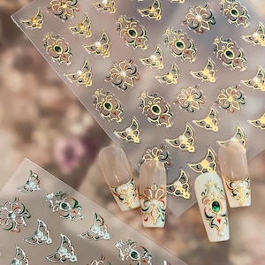 Può includere: Primo piano di adesivi per nail art con disegni floreali e geometrici dorati, verdi e rossi. Diverse unghie sono decorate con motivi simili, con perle e pietre preziose. Gli adesivi sono su un foglio trasparente.