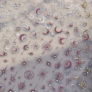 May include: A sheet of iridescent nail art stickers featuring celestial designs. The stickers include moons, stars, suns, and other cosmic elements in silver, red, and purple hues. Some designs have embedded rhinestones for added sparkle.
