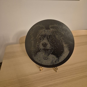 May include: A round, black decorative piece with a pattern of small, evenly spaced holes, creating a design. The piece is displayed on a wooden stand. The background includes a framed artwork and a light-colored wooden table.