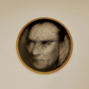 Puede incluir: Arte mural circular que presenta un retrato creado con hilo negro. La obra de arte representa el rostro de un hombre, con el hilo formando los rasgos sobre un fondo oscuro. El marco es de color marrón claro.