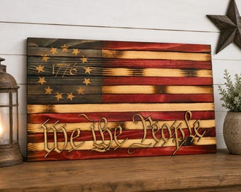 May include: A rectangular wooden wall sign featuring a burnt-wood American flag design with thirteen stars arranged in a circle around the year 1776. The phrase We the People is carved in a decorative script font across the lower red and white stripes.