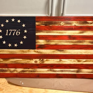 May include: Wooden American flag with a distressed finish. The flag features red and white stripes, a blue canton with a circle of stars, and the year "1776" in the center. The wood grain is visible, adding to the rustic aesthetic.