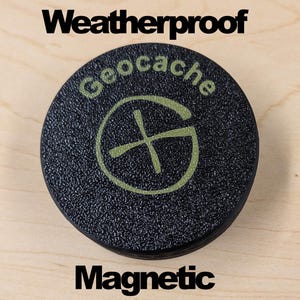 May include: A round, black, textured magnetic geocache container featuring the word Geocache in light green text above a circular green logo with a cross symbol. The words Weatherproof and Magnetic are printed in black above and below the object.