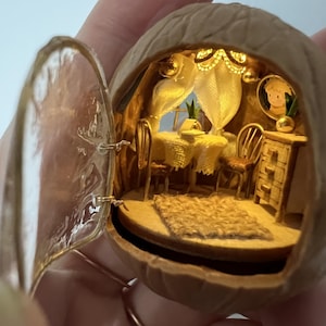 Może przedstawiać: Miniaturowa scena domku dla lalek wewnątrz skorupy orzecha włoskiego. Wnętrze zawiera mały stół, krzesła, dywan i oprawiony obraz, wszystko oświetlone ciepłym światłem. Skorupa jest otwarta, ukazując szczegółowe rzemiosło małego pokoju.