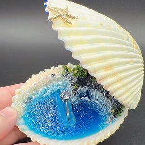 May include: A ring holder crafted from a large, open white seashell featuring a miniature diorama of a blue ocean cove with rocky cliffs and green foliage. A gold-toned starfish is attached to the top shell, and a silver ring is centered.