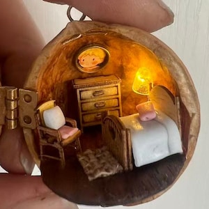 May include: A miniature bedroom scene housed within a walnut shell. The interior includes a bed, chair, dresser, and a framed picture. A small lamp provides warm lighting. The shell is hinged with brass hardware, creating a unique, handcrafted piece.