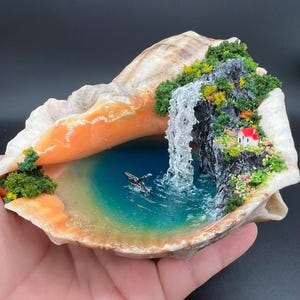 Op de afbeelding: Miniatuur oceaan diorama in een grote schelp. De scène omvat een waterval, een klein huisje met een rood dak en een walvis. Het water is turquoise en de schelp heeft oranje en witte tinten.