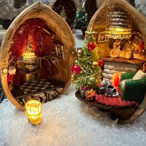 May include: Miniature Christmas scene inside a walnut shell. One shell features a decorated Christmas tree, presents, and a cat. Another shell has a small room with a lamp and a checkered floor. A lit candle sits in front.