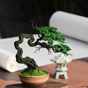 Op de afbeelding: Een miniatuur bonsai-boom met een gedraaide stam en heldergroene bladeren. De boom staat in een kleine, ronde, bruin-oranje pot. Een kleine stenen lantaarn staat ernaast, wat een serene, zen-achtige scène creëert.