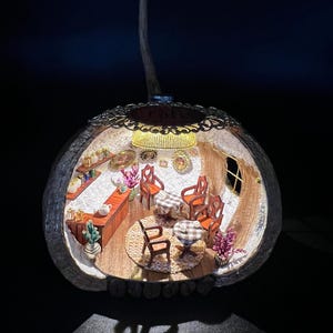 May include: A miniature cafe diorama inside a decorative, illuminated shell. The interior features a bar, tables, chairs, and decorative plants. The shell is adorned with a delicate, lacy trim and a hanging loop.