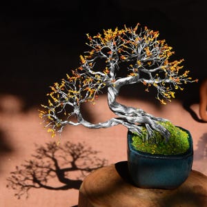 Op de afbeelding: Een handgemaakte bonsai boomsculptuur met een zilverkleurige stam en takken, versierd met gele en oranje bladeren. De miniatuurboom staat in een teal-kleurige pot gevuld met groen mos, geplaatst op een houten voet. De schaduw van de boom is zichtbaar.