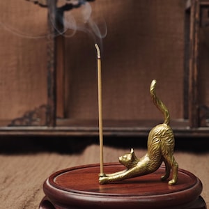 Peut inclure: Un porte-encens doré en forme de chat en position d'étirement, tenant un bâton d'encens allumé. Le support est posé sur un socle rond en bois foncé. Des volutes de fumée s'élèvent de l'encens, créant une atmosphère apaisante.