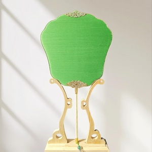 Può includere: Un ventaglio decorativo verde con bordo smerlato, posto su un supporto in legno chiaro. Il ventaglio presenta dettagli in filigrana color oro e un manico abbinato. Il supporto ha una base rettangolare e supporti curvi.