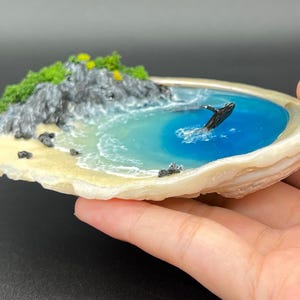 Pode incluir: Uma cena oceânica em miniatura numa concha. A cena apresenta uma baleia saltando em água azul, uma praia arenosa e uma falésia rochosa com folhagem verde. A concha é esbranquiçada com uma superfície texturizada.