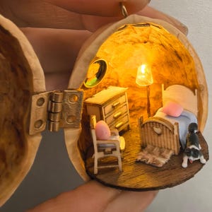 Könnte beinhalten: Eine Miniatur-Schlafzimmerszene in einer Walnussschale, beleuchtet von einer kleinen Lampe. Die Szene umfasst ein Bett, eine Kommode, einen Stuhl und eine kleine Hundefigur. Die Walnussschale ist geöffnet und zeigt das detaillierte Innere.