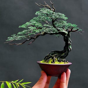 May include: A handcrafted bonsai tree sculpture. The tree features a twisted, dark brown trunk and branches, with vibrant green wire foliage. The bonsai sits in a small, reddish-brown ceramic pot filled with moss. The sculpture is held by a hand.