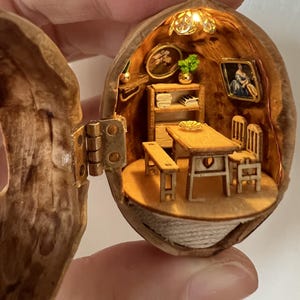 May include: A miniature dollhouse scene inside a walnut shell. The interior features a small table, chairs, a bench, a bookshelf, and framed artwork. The scene is illuminated by a small light fixture, creating a warm, inviting ambiance.
