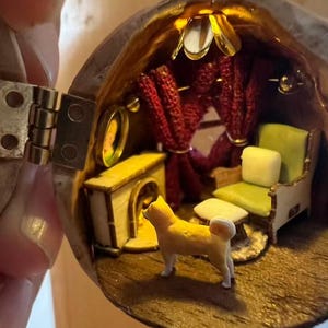 May include: A miniature diorama inside a walnut shell, featuring a tiny yellow dog standing in a furnished room. The room includes a green armchair, red curtains, and a small fireplace, all illuminated by a warm light.