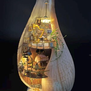 Pode incluir: Diorama em miniatura de um quarto dentro de uma concha. O interior apresenta uma harpa dourada, uma cama e uma escada em espiral. A concha é iluminada com iluminação quente, criando uma atmosfera acolhedora e caprichosa. A base é de um material branco texturizado.