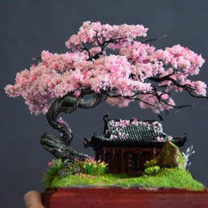 Op de afbeelding: Miniatuur bonsai boom met een klein traditioneel Aziatisch gebouw. De boom heeft een gedraaide stam en is bedekt met roze bloesems. Het gebouw heeft een donkergroen dak en is omgeven door groen gras en kleurrijke bloemen. De basis is donkerrood.