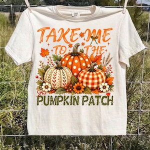Peut inclure: Un t-shirt à manches courtes de couleur crème avec une impression graphique de trois citrouilles aux motifs à pois, floraux et à carreaux. Le texte "TAKE ME TO THE PUMPKIN PATCH" est imprimé en orange et vert, entouré de feuilles d'automne.