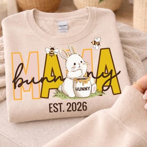 Peut inclure: Sweat-shirt beige avec un motif graphique représentant les mots "Mama Bunny" en écriture noire et en lettres majuscules jaunes. Un lapin blanc avec un pot de miel et deux abeilles sont également représentés. Le texte "EST. 2026" est en dessous.