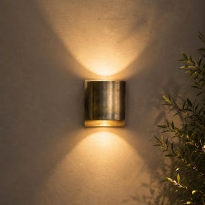 May include: A cylindrical wall sconce with a brushed brass finish. The sconce emits a warm, inviting light, casting a soft glow on the surrounding wall. The fixture is mounted on a neutral-toned wall, with a hint of greenery in the corner.