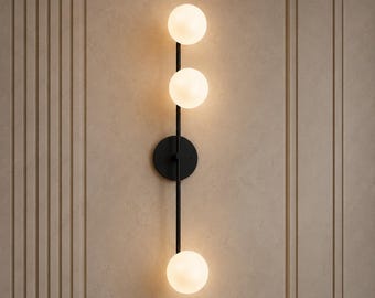 Puede incluir: Aplique de pared moderno con estructura de metal negro y tres bombillas esféricas de vidrio esmerilado. El aplique está montado en una pared beige con paneles verticales, creando un diseño minimalista y elegante.