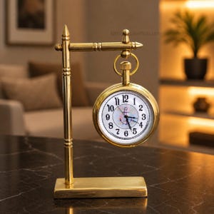May include: A brass-coloured, vintage-style desk clock. The clock face is white with black numerals and hands, and a nautical-themed design. The clock hangs from a decorative stand with a rectangular base.