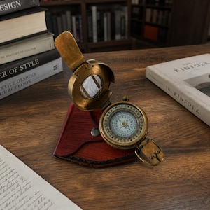 May include: A vintage brass compass in a red leather case. The compass dial is detailed and the lid is open. The compass is on a wooden surface. The case is a deep red colour. The compass is a classic navigational tool.
