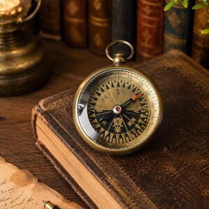 May include: A brass compass with a glass face, positioned on a weathered leather-bound book. The compass needle indicates north. An oil lamp and antique books are in the background, evoking a sense of history and exploration.