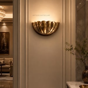 Könnte beinhalten: Goldfarbene Wandleuchte mit muschelförmigem Design an einer beigefarbenen Wand. Die Leuchte ist beleuchtet und erzeugt einen warmen Schein. Der Text "ANTIQUELIGHTGELLARY" ist über der Leuchte zu sehen. Es ist eine dekorative Leuchte.