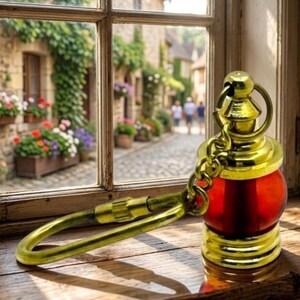 Puede incluir: Un llavero decorativo de estilo antiguo con un orbe de vidrio rojo y detalles metálicos dorados. El llavero tiene un cierre grande y curvo y una pequeña cadena. El fondo muestra una ventana con vistas a una calle europea.
