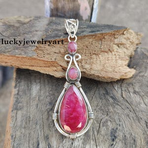 Pode incluir: Pingente de prata com uma grande pedra de rubi em forma de gota. Dois rubis ovais menores estão definidos acima da pedra principal. O pingente tem um design decorativo e ondulado e é exibido em uma superfície de madeira. O texto "luckyjewelryart" é visível.