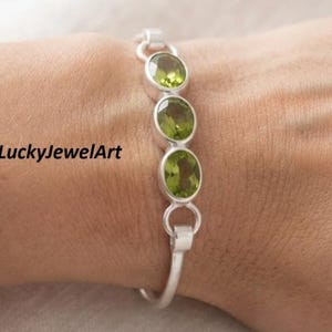 Op de afbeelding: Een zilveren armband met drie ovaalvormige, gefacetteerde groene peridoot edelstenen in gladde metalen zettingen, verbonden door cirkelvormige zilveren schakels. De tekst LuckyJewelArt is zichtbaar aan de linkerkant van de afbeelding.