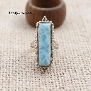 May include: A rectangular larimar stone ring set in a silver frame with a twisted rope border. The stone features a light blue marbled pattern. The text LuckyJewelArt is visible in the upper left corner of the image, positioned above the ring.