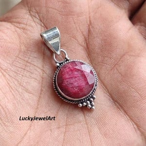Pode incluir: Pingente de prata com uma grande pedra preciosa de rubi oval e facetada. O rubi é de cor vermelha profunda e está engastado em uma moldura decorativa de prata. O pingente tem uma argola para prender a uma corrente. O texto "LuckyJewelArt" é visível.