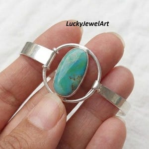 Könnte beinhalten: Ein silbernes Armband mit einem zentralen ovalen Türkisstein mit blauer und grüner Marmorierung. Der Stein ist in einen kreisförmigen Silberrahmen gefasst, der mit den beiden Seiten des Armbands verbunden ist. Der Text LuckyJewelArt ist sichtbar.