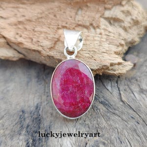 Pode incluir: Um pingente de prata com uma pedra preciosa de rubi vermelho escuro oval. O rubi é colocado em uma moldura de prata e tem uma superfície polida. O pingente tem uma argola simples para uma corrente. O texto "luckyjewelryart" está na parte inferior.