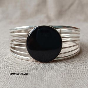 Può includere: Un bracciale a polsino argentato con una grande pietra nera rotonda al centro. Il bracciale ha più bande parallele. Il gioiello è esposto su una superficie di colore neutro. Il testo "LuckyJewelArt" è visibile.