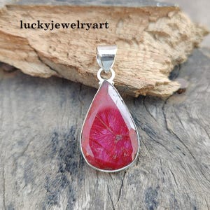 Pode incluir: Um pingente em forma de gota com uma pedra preciosa vermelha vibrante emoldurada em uma estrutura de metal prateado. A pedra preciosa tem um padrão texturizado único. O pingente inclui uma argola prateada para uma corrente. O texto "luckyjewelryart" é visível.