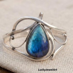May include: A silver cuff bracelet featuring a teardrop-shaped labradorite gemstone with blue and purple iridescence. The metal band has a swirling, open-work design with thin, curved lines and small spherical accents. The text LuckyJewelArt is visible.