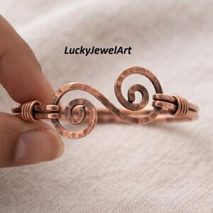 May include: A hammered copper cuff bracelet featuring a central double spiral design. The metal surface has a textured, dimpled finish with wire-wrapped details at the ends. The text LuckyJewelArt is visible above the copper bracelet.