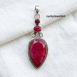 May include: A silver pendant featuring a large pear-shaped faceted red ruby at the base, topped by a smaller oval ruby and a tiny round ruby. The metal setting includes decorative scrollwork and a bail at the top. The text LuckyJewelArt is visible.