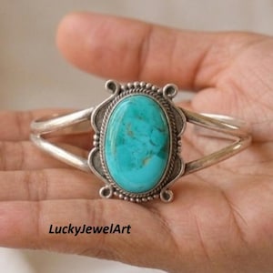 May include: A silver cuff bracelet featuring a large, oval-shaped turquoise stone at the centre. The stone is framed by a twisted rope border and decorative silver scrollwork. The band is split into two thin, polished silver arms on each side.