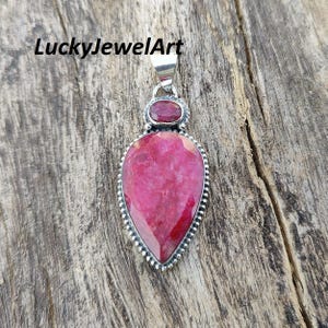 Pode incluir: Pingente de prata com uma grande pedra rosa em forma de gota. Uma pedra rosa ovalada menor está localizada acima da pedra maior. O pingente tem uma borda texturizada e uma argola de prata. O texto "LuckyJewelArt" está no canto superior esquerdo.