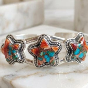 Könnte beinhalten: Ein silbernes Armband mit drei sternförmigen Einlagen. Jeder Stern enthält ein Mosaik aus leuchtend türkisfarbenen und orangeroten Steinfragmenten, die von einer gedrehten Silberkordel umrandet sind. Das Metallband ist poliert.