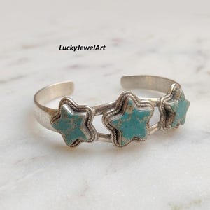 May include: A silver cuff bracelet featuring three star-shaped inlays of turquoise stone with brown matrix veining. The metal band has a twisted rope texture bordering each star. The text LuckyJewelArt is visible above the bracelet.