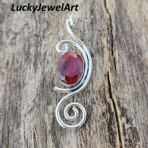 Pode incluir: Pingente de prata com uma grande pedra preciosa ovalada vermelha escura. O pingente tem um design artístico em espiral. O fundo é uma superfície de madeira. O texto "LuckyJewelArt" está no canto superior esquerdo.