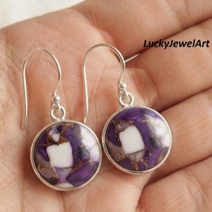 May include: A pair of drop earrings featuring round, bezel-set stones with a mosaic pattern of deep purple, white, and copper-colored veins. The earrings have silver-toned metal hooks and circular settings. The text LuckyJewelArt is visible.